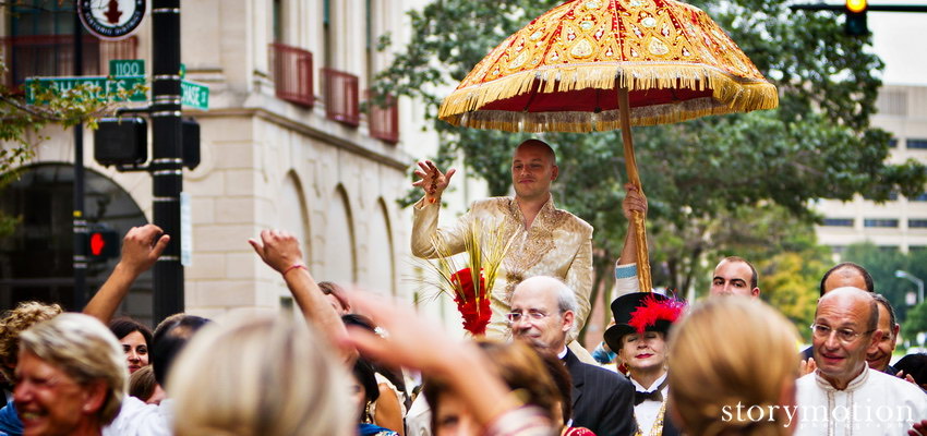 A groom rides in a baraat procession under a red and gold umbrella