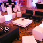 A modern lounge area with black and white furniture, including sofas, armchairs, a low table, and cube seats, set on a carpet with a leaf pattern. Dining tables are visible in the background.