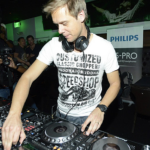DJ wearing headphones is operating audio equipment in a dimly lit venue. Several people and a photographer are in the background. A Philips promotional banner is visible.
