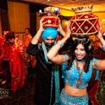 A group of people in colorful attire dance at an Indian wedding, with two individuals carrying traditional pots on their heads.