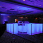 A dimly lit banquet hall with blue neon lights illuminating a U-shaped bar area. Round tables with black tablecloths and white chairs are visible in the background.