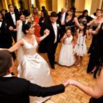 A bride and groom, along with their guests, hold hands in a circle while dancing at a wedding reception.