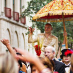 A man in traditional attire holds a golden umbrella while being lifted through a crowd on a city street during a public event.