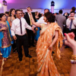 A group of people in traditional attire dancing and celebrating at an indoor event. A woman in an orange sari is in the foreground with her arm raised.