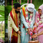 A group of people in traditional attire participate in a ceremonial ritual, placing hands over a vessel. Floral garlands and pink decorations are present in the background.