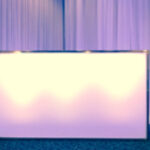 A brightly lit, rectangular table with a white tablecloth stands against a backdrop of flowing purple curtains.