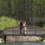 A couple dressed in formal attire stand close together on a small wooden bridge over a creek, surrounded by lush greenery and trees.