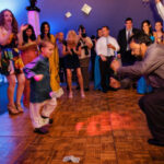 A young child and a man dance on a wooden floor surrounded by a cheering crowd at an event; dollar bills are scattered on the floor.