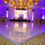 A spacious, elegantly decorated banquet hall with purple lighting. The room features a large dance floor, neatly arranged tables with white chairs, and a stage with a decorative backdrop.