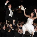A bride and groom are lifted into the air on chairs by a group of people at a wedding reception, smiling and holding hands. The crowd around them is celebrating in a dimly lit venue.
