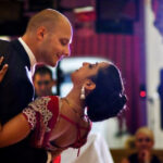 A couple shares a dance at what appears to be a wedding reception, the woman wearing a red and silver traditional outfit, and the man in a dark suit. The background is lit with warm, festive lighting.