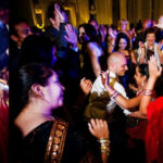 A group of people are dancing and celebrating together indoors. Some women are dressed in colorful traditional clothing. A man in the center is dancing energetically with the crowd around him.