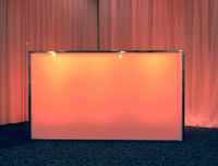 A large rectangular pink partition is illuminated by overhead lights. It stands in front of pink draped curtains in a dimly lit room.
