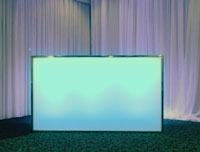 An illuminated rectangular panel in front of floor-to-ceiling white curtains. The panel emits a soft blue glow in a dimly lit room.